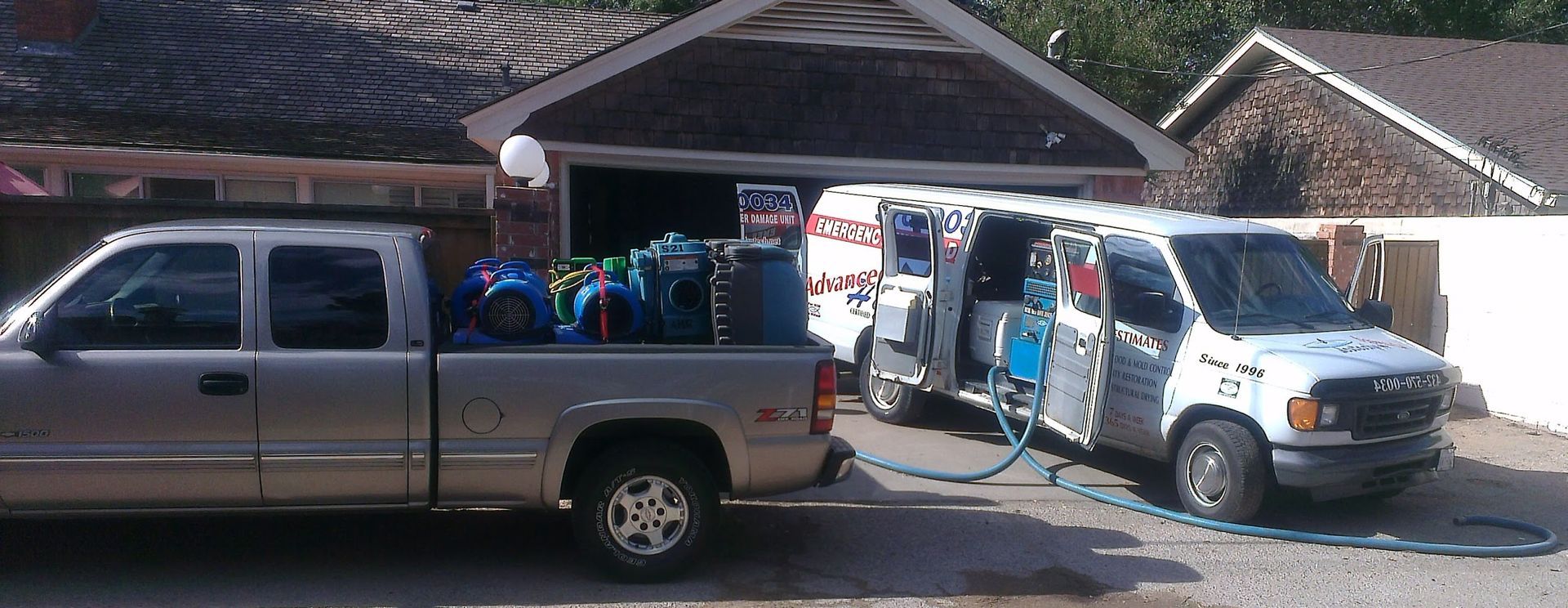 A silver pickup truck parked next to a white service van with the doors open in front of a garage.