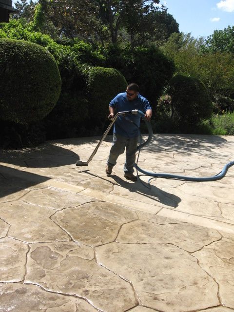 Man using vacuum cleaner on a wet, stone-patterned patio, surrounded by greenery. Sunny day.