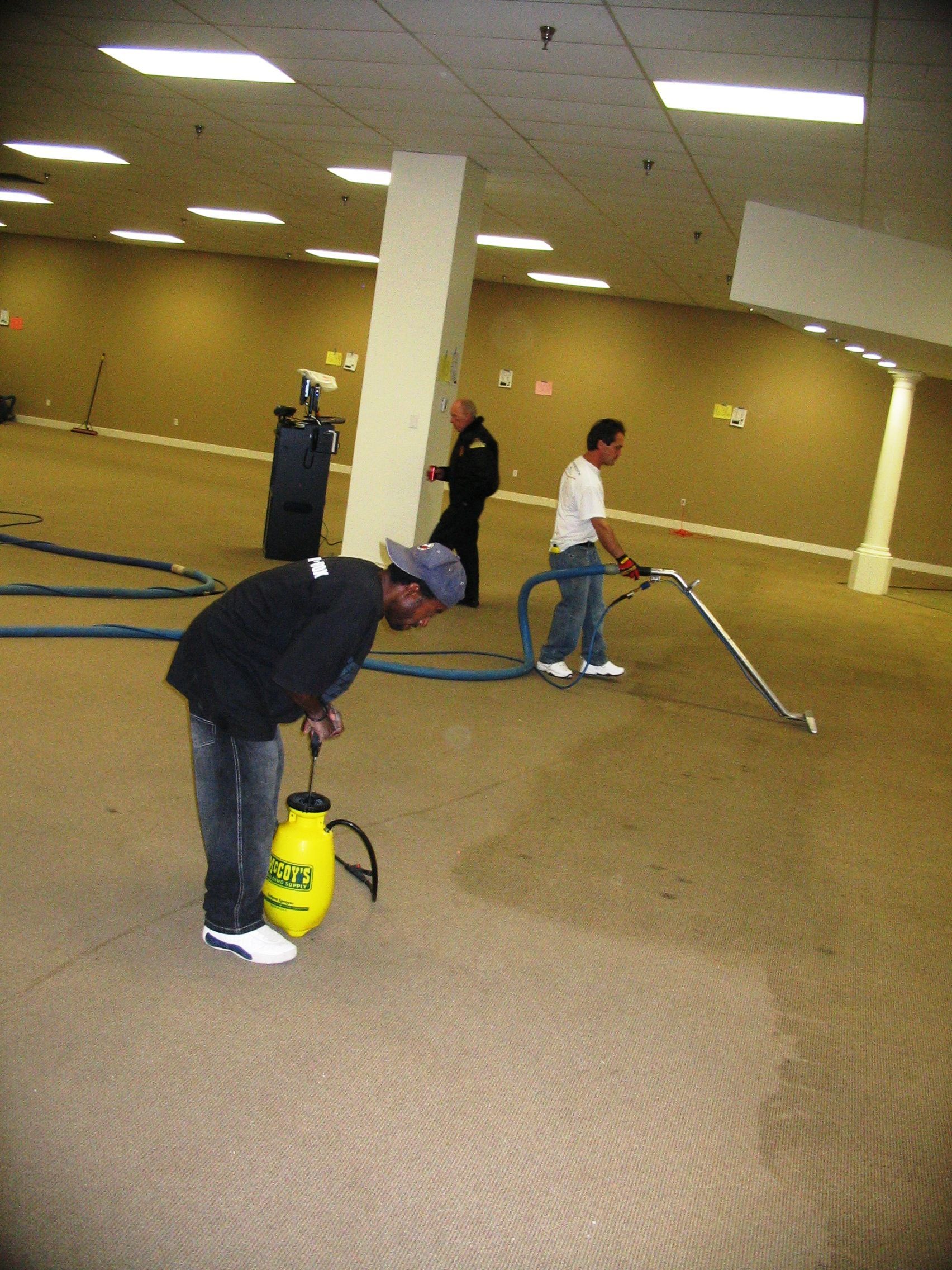 Men cleaning carpet in a large room; one spraying, one operating a cleaning machine, and one observing.