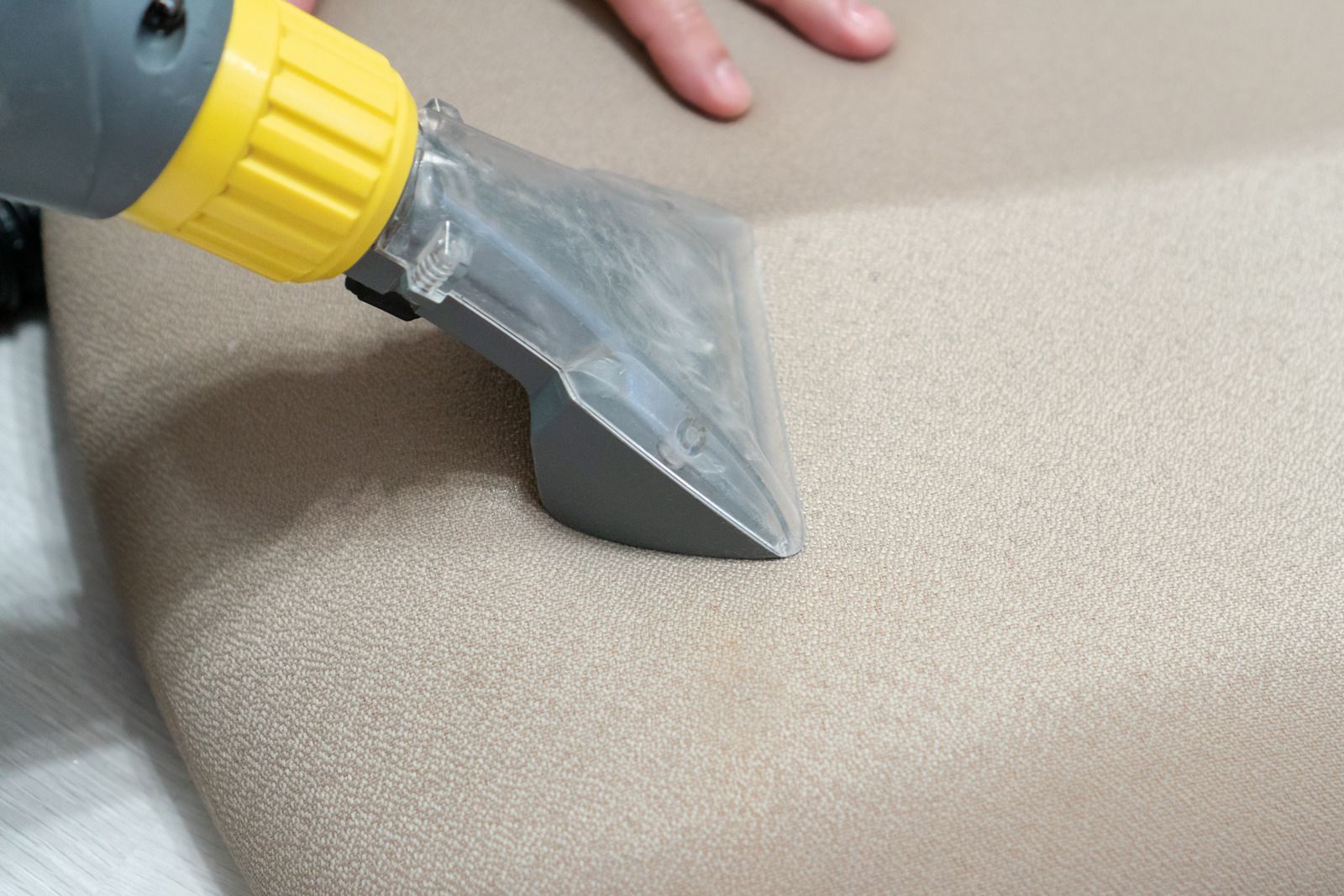 A person cleaning a beige upholstered seat with a handheld upholstery cleaner.