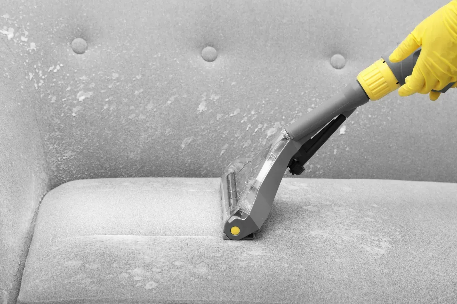 Person in yellow glove vacuums a light gray sofa with cleaning residue.