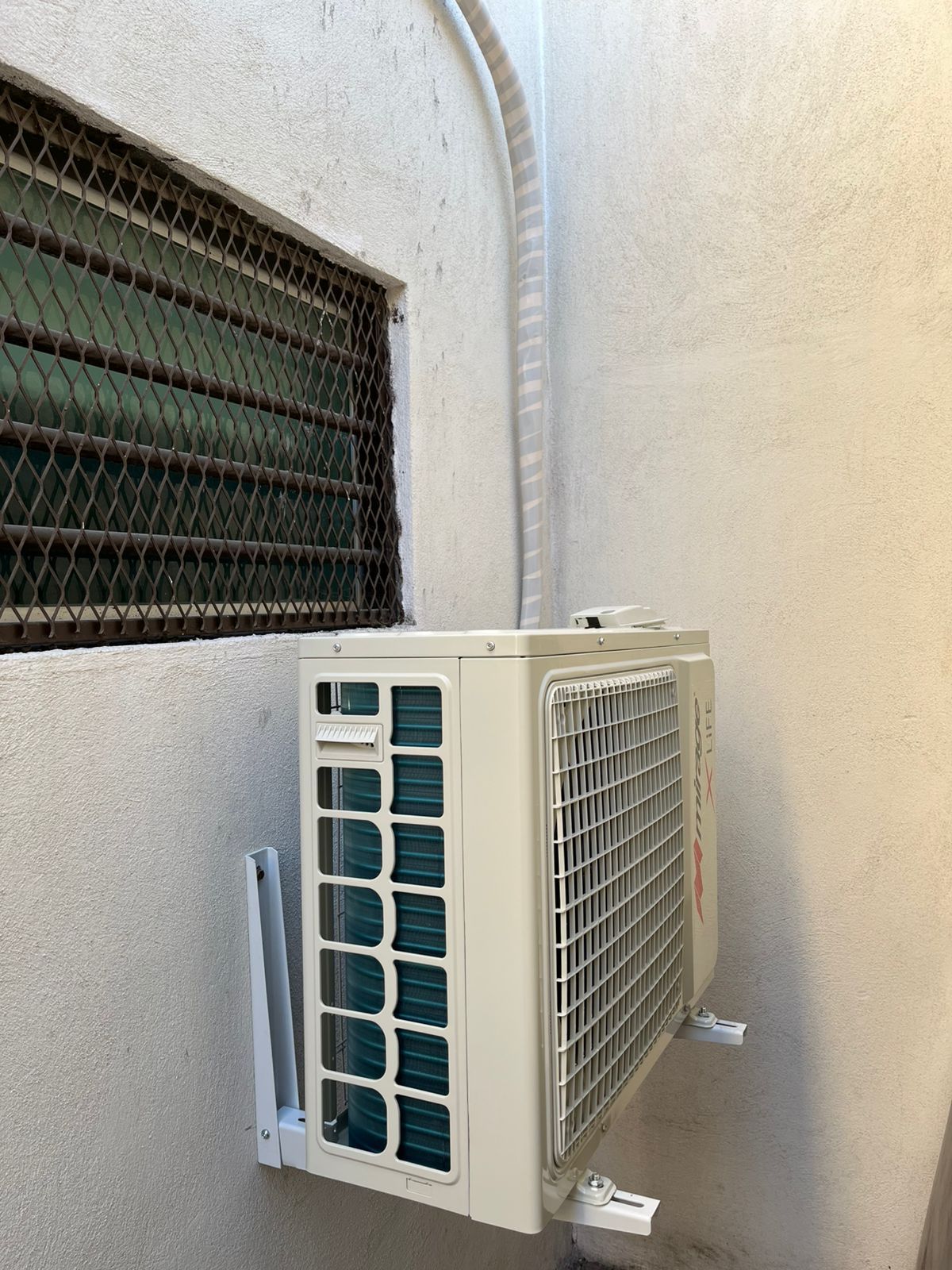 Una unidad condensadora de aire acondicionado montada en una pared exterior de color claro junto a una ventana con una reja de seguridad metálica.
