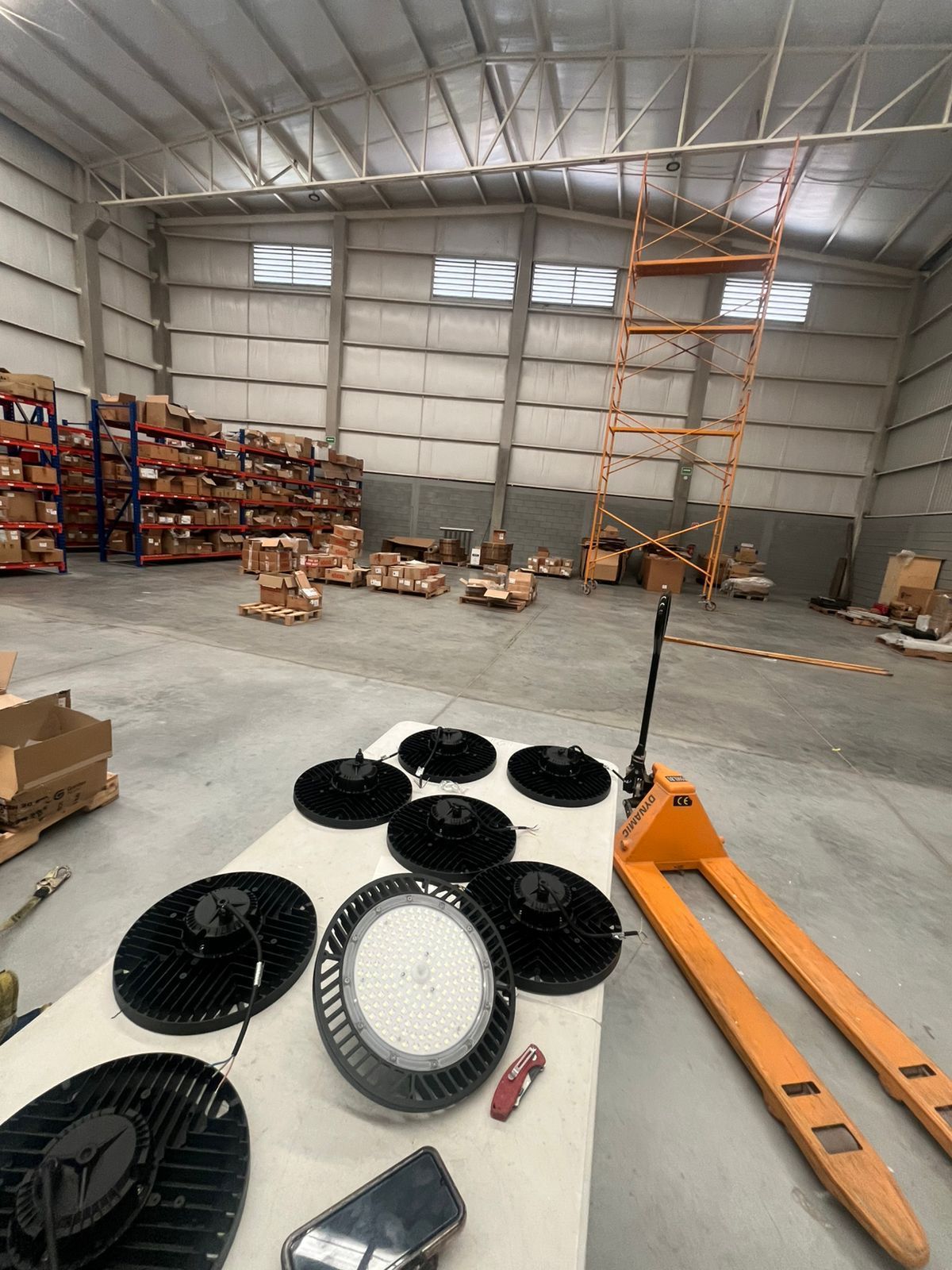 Varias luminarias industriales circulares se encuentran sobre una mesa blanca dentro de un almacén grande y escaso con estanterías de metal.