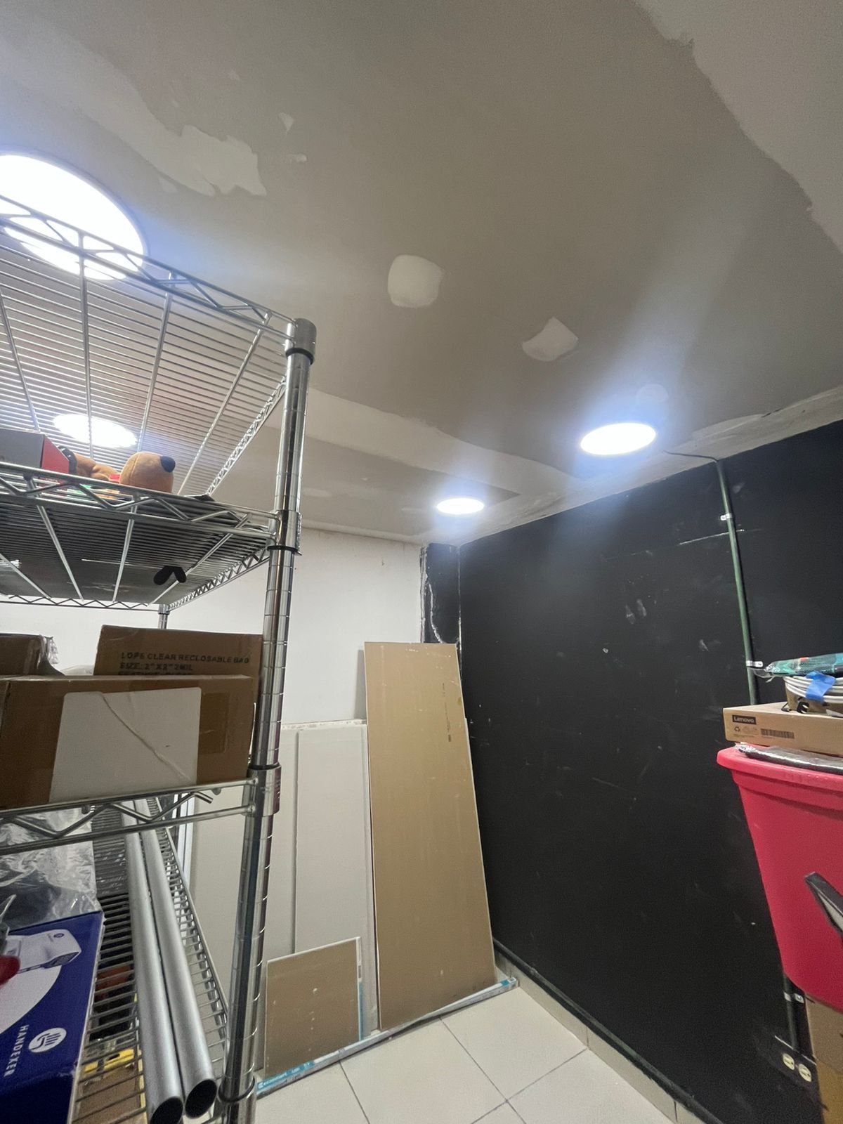 Área de almacenamiento con estanterías metálicas, cajas y paneles de pared, iluminada por brillantes luces circulares en el techo.