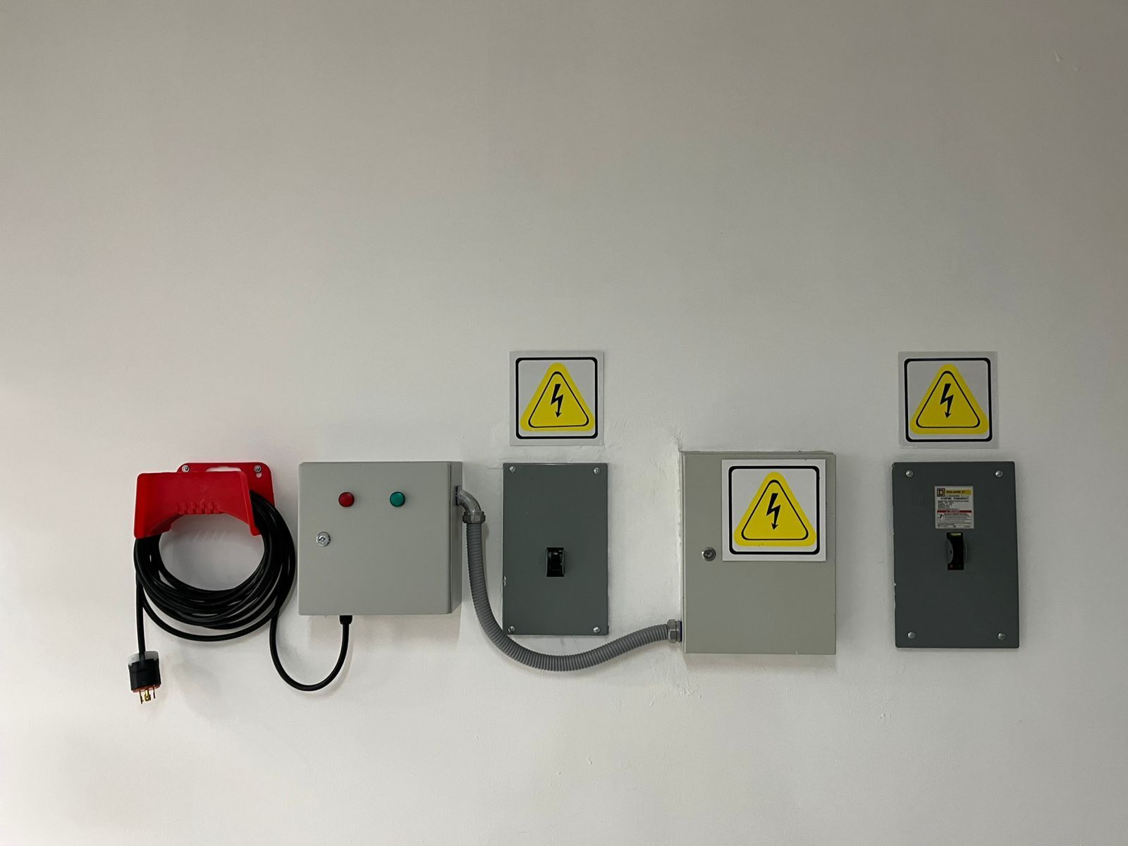Equipo eléctrico montado en una pared blanca, que incluye un cable enrollado, una caja de control y dos paneles de disyuntores.