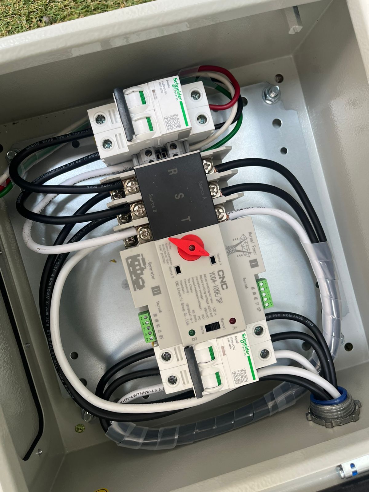 Un gabinete eléctrico abierto que contiene un interruptor de transferencia blanco con un dial rojo, conectado a dos disyuntores blancos.
