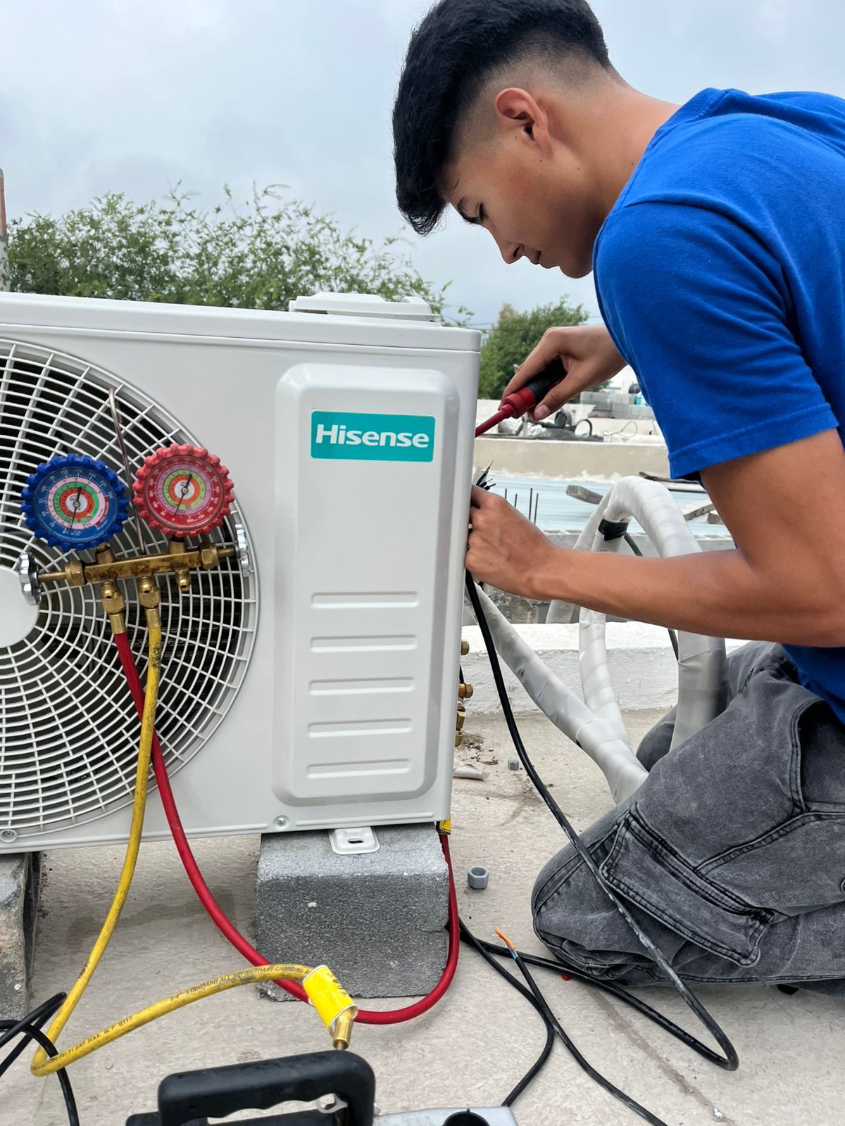 Un técnico con una camisa azul trabaja en una unidad de aire acondicionado exterior Hisense utilizando un juego de manómetros y un destornillador.