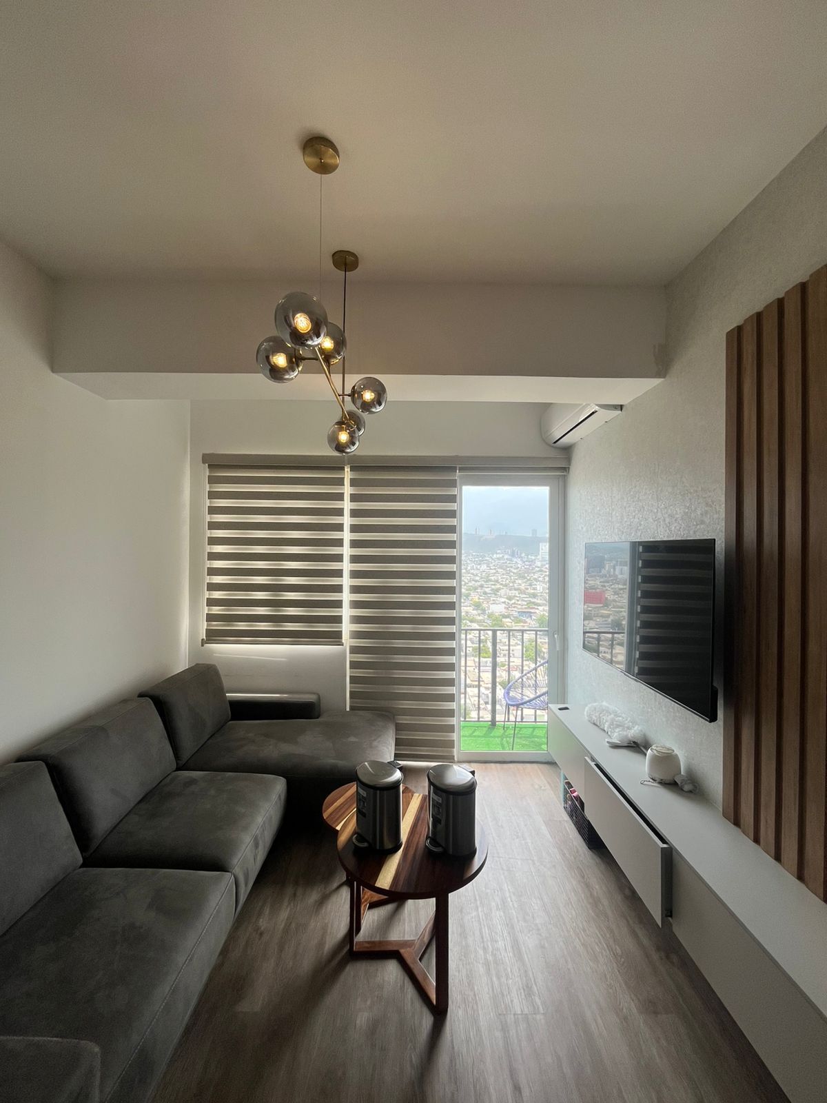 Una sala de estar moderna con un sofá seccional gris, una pequeña mesa de café de madera, un televisor montado en la pared y una lámpara de araña de cristal.