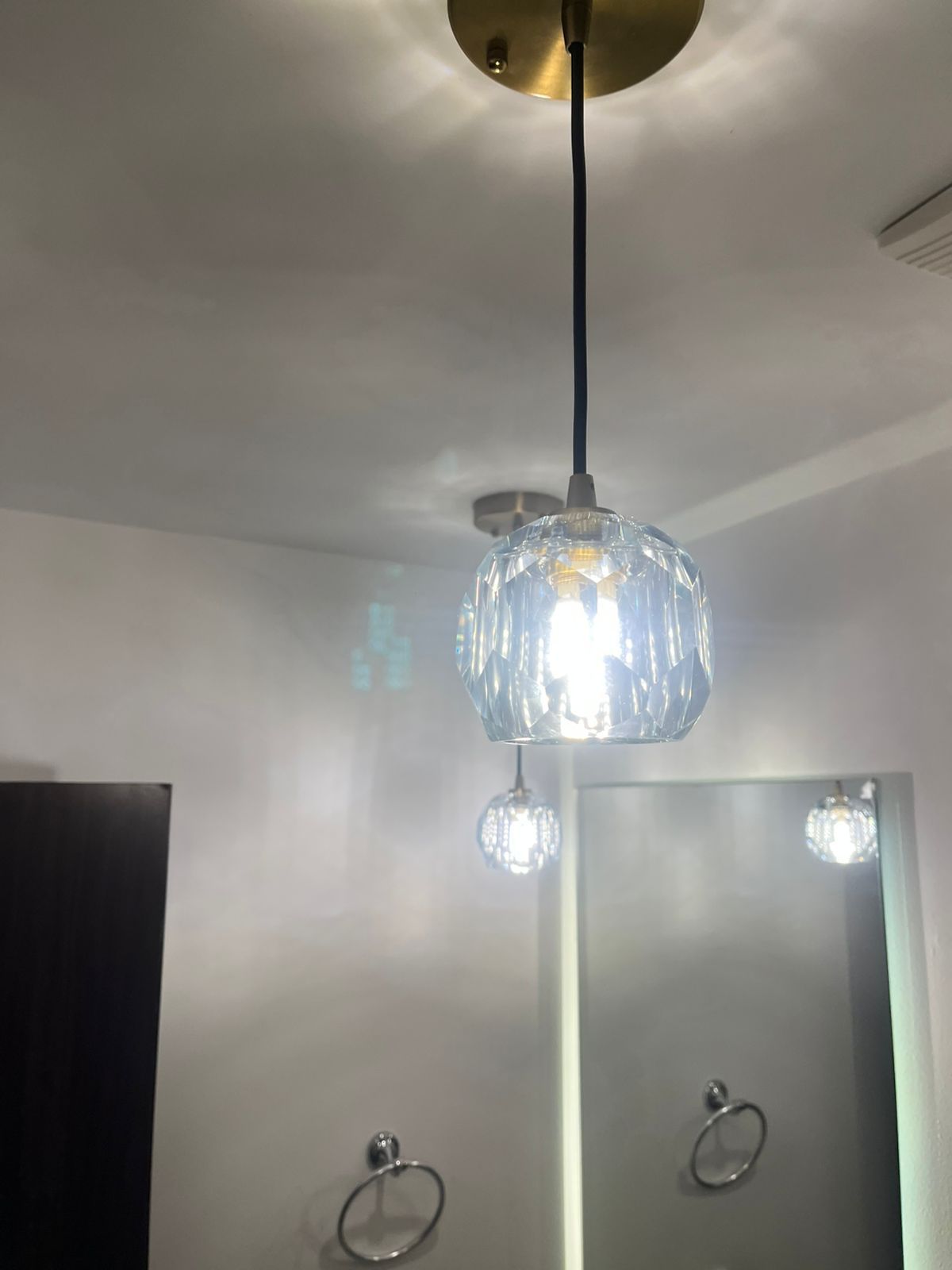 Una lámpara colgante de vidrio iluminada y multifacética cuelga de un soporte de techo de latón en una habitación con una pared blanca y un espejo de tocador.