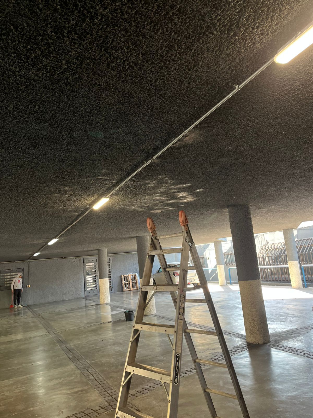 Una escalera de mano se encuentra en un amplio estacionamiento abierto con techos oscuros, pilares de hormigón y luces de tira brillantes.