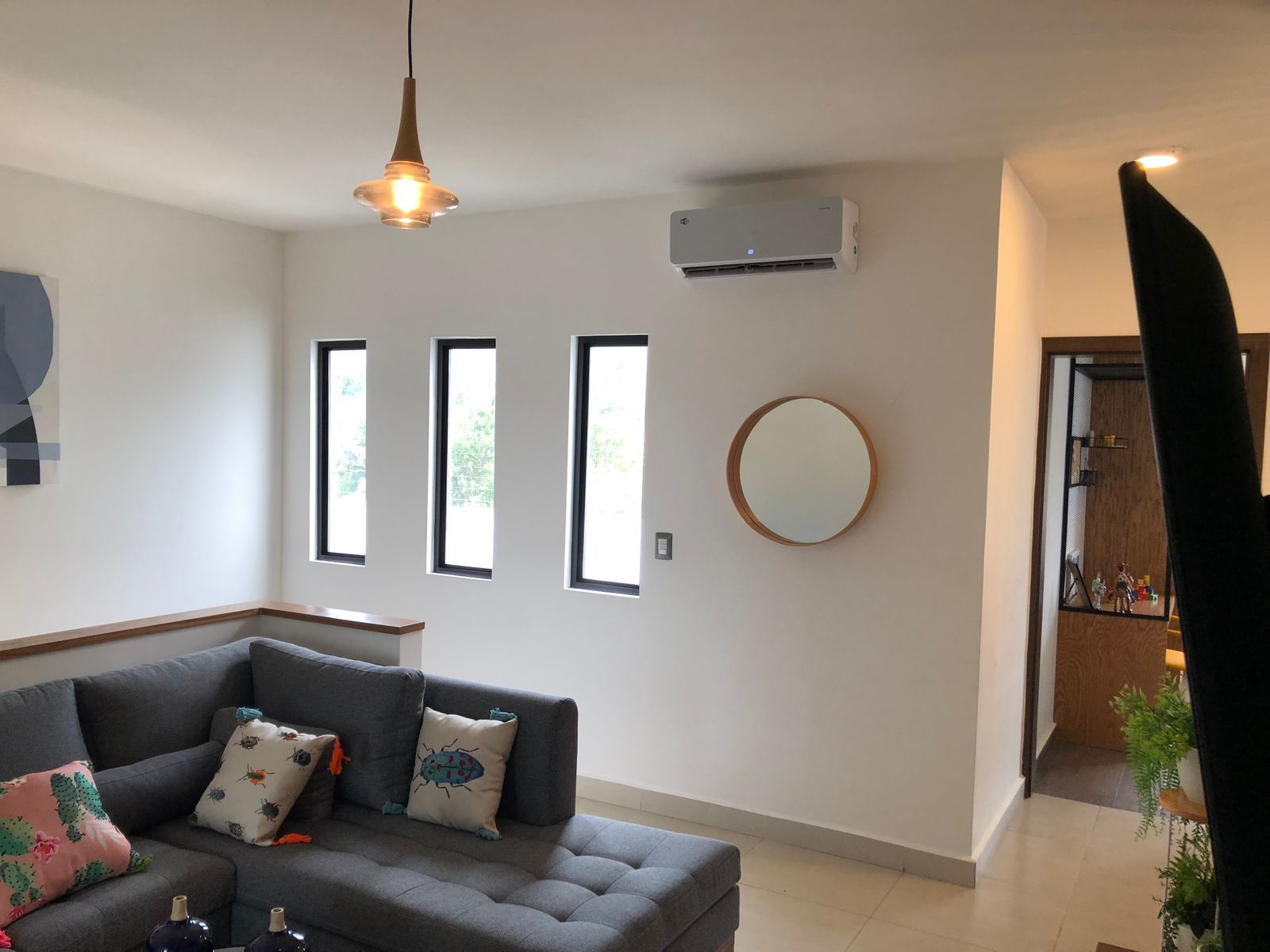 Una sala de estar moderna con un sofá seccional gris, tres ventanas estrechas, un espejo redondo y un aire acondicionado montado en la pared.