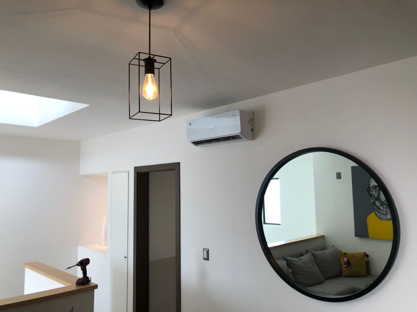 Una habitación blanca con una lámpara colgante geométrica, un aire acondicionado montado en la pared y un gran espejo redondo con marco negro.