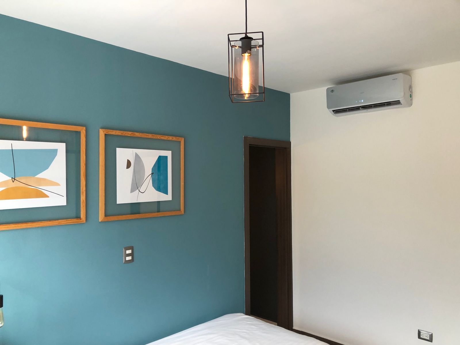 Una pared de dormitorio con pintura verde azulado, dos impresiones abstractas enmarcadas, una lámpara colgante moderna y un aire acondicionado montado en la pared.