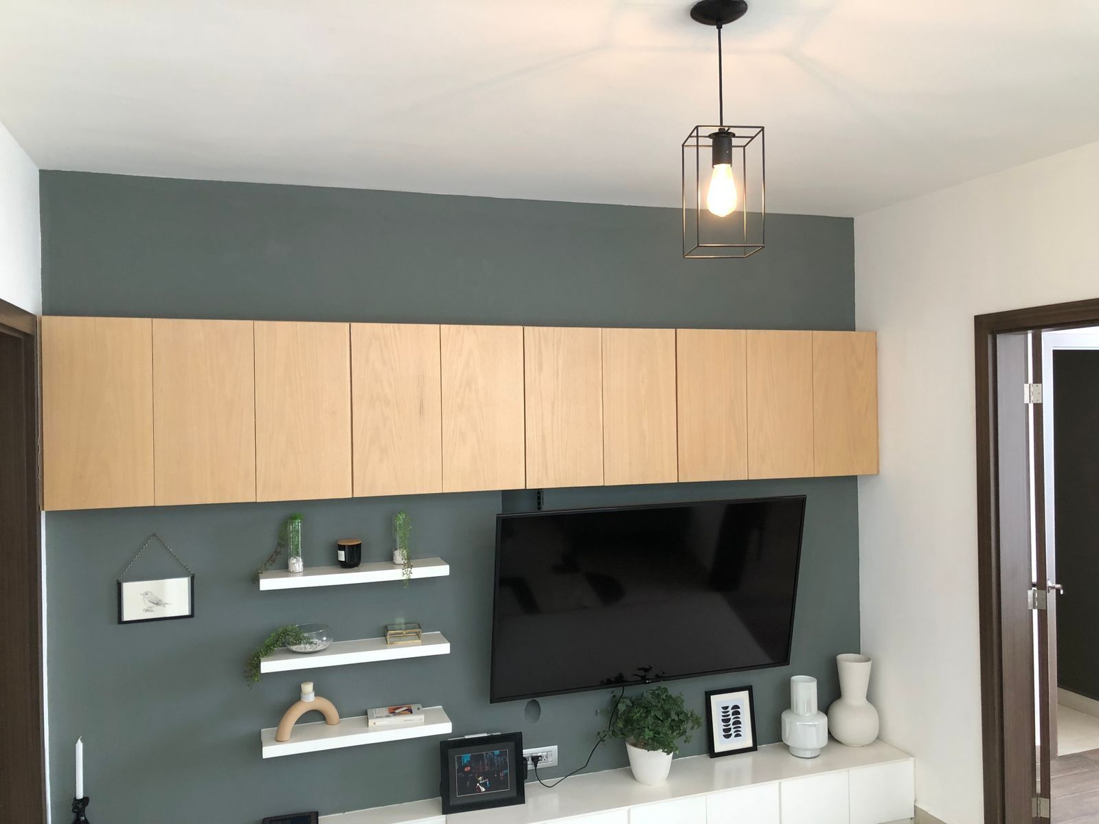 Una pared de sala de estar gris con un mueble flotante de madera, un televisor montado en la pared, tres estantes con decoración y una lámpara colgante.