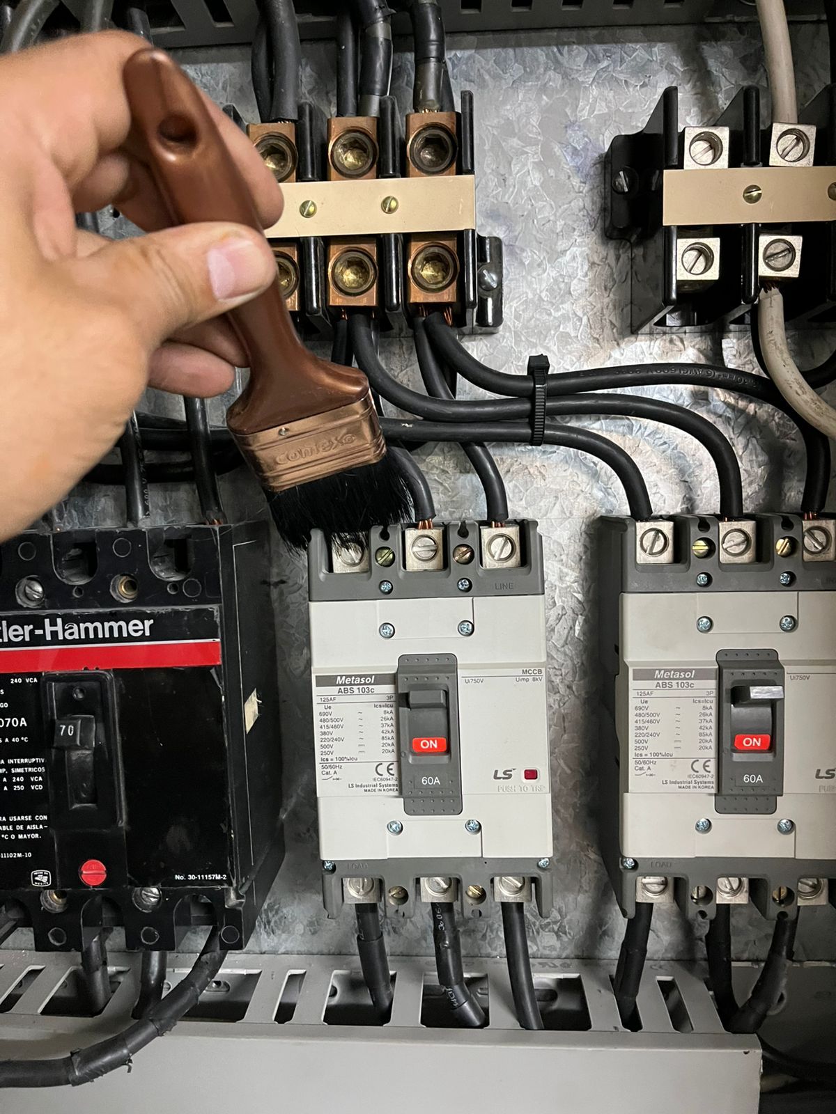 Una mano utiliza un cepillo pequeño para limpiar el polvo de los interruptores eléctricos dentro de un panel de control.