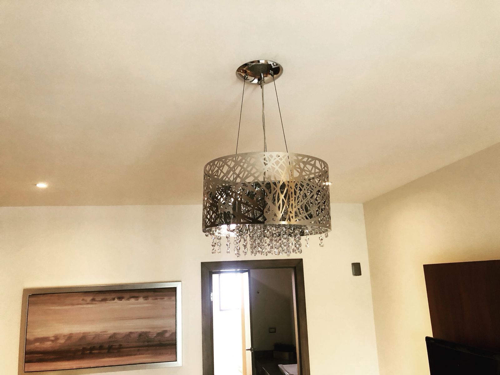 Una moderna lámpara colgante estilo tambor con un patrón de filigrana metálica y detalles de cristal colgantes en una habitación.