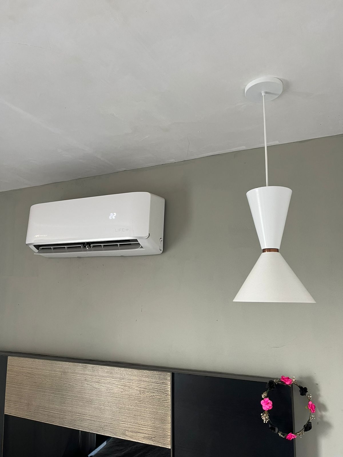 Una unidad de aire acondicionado montada en la pared de color blanco junto a una moderna lámpara colgante blanca en forma de cono en una habitación.