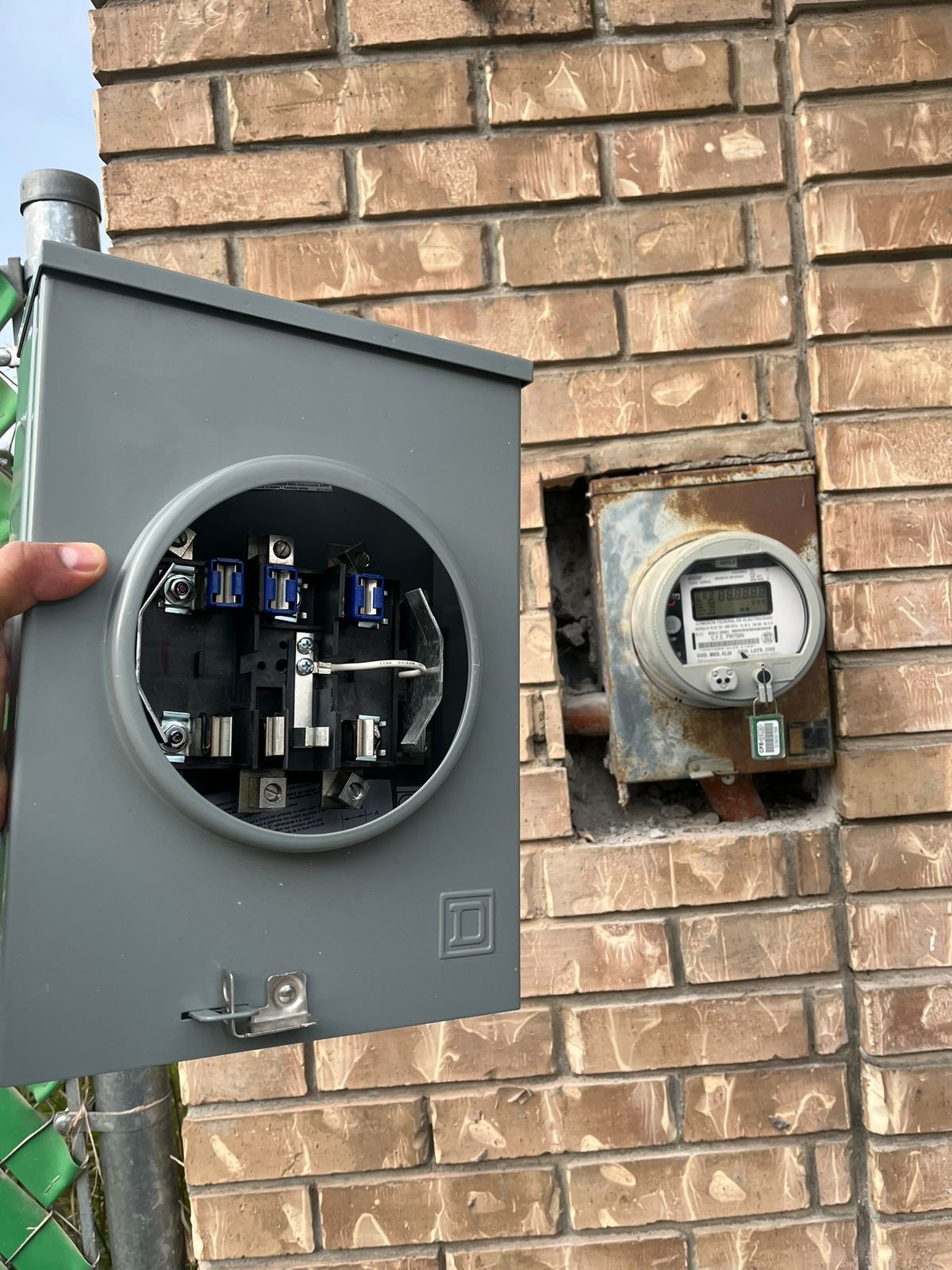 Una persona sostiene una caja de medidor eléctrico de metal gris abierta frente a un medidor de servicios públicos digital montado en una pared de ladrillos.
