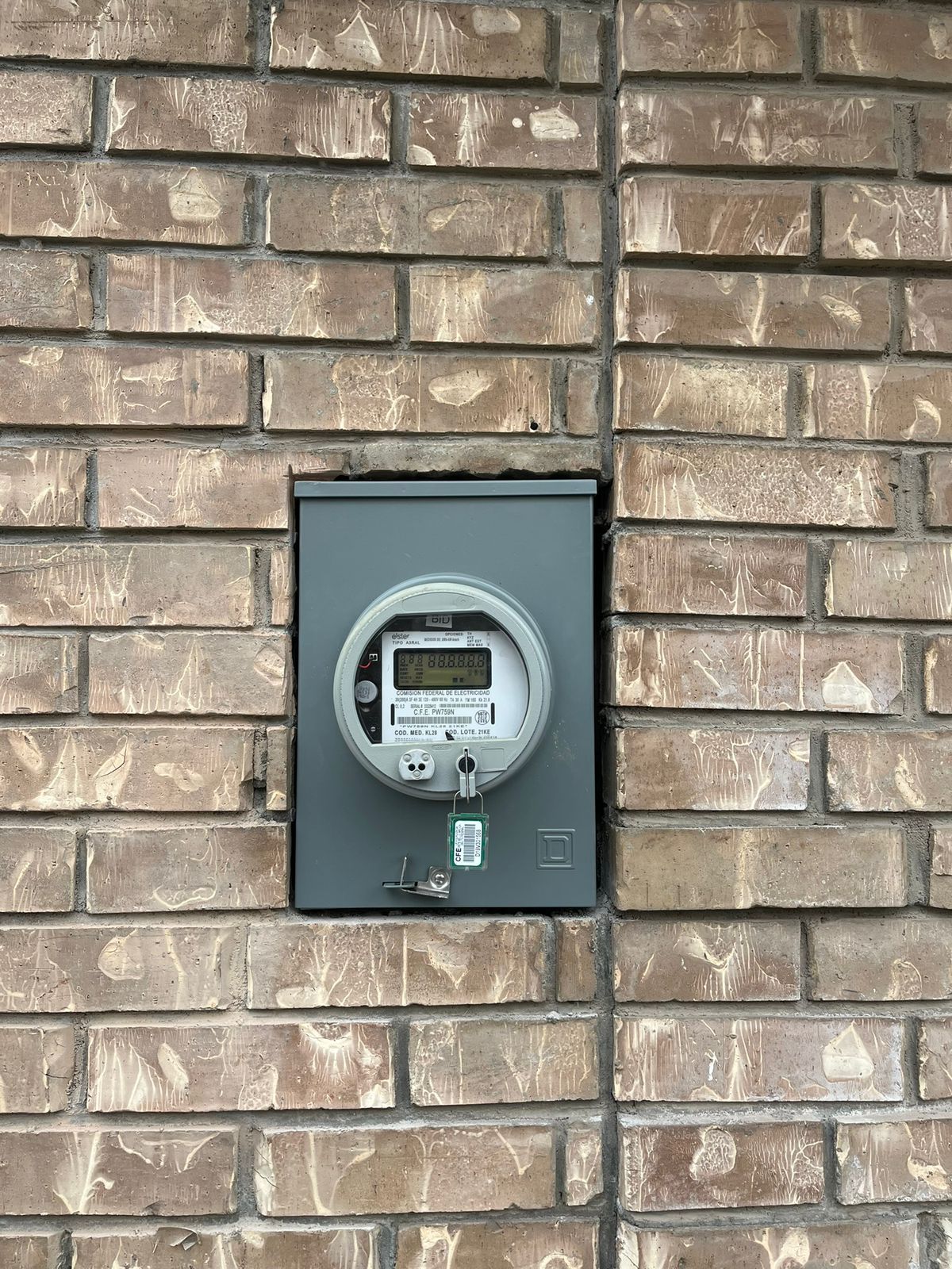 Un medidor eléctrico digital montado en una caja de metal gris sobre una pared de ladrillos color canela.