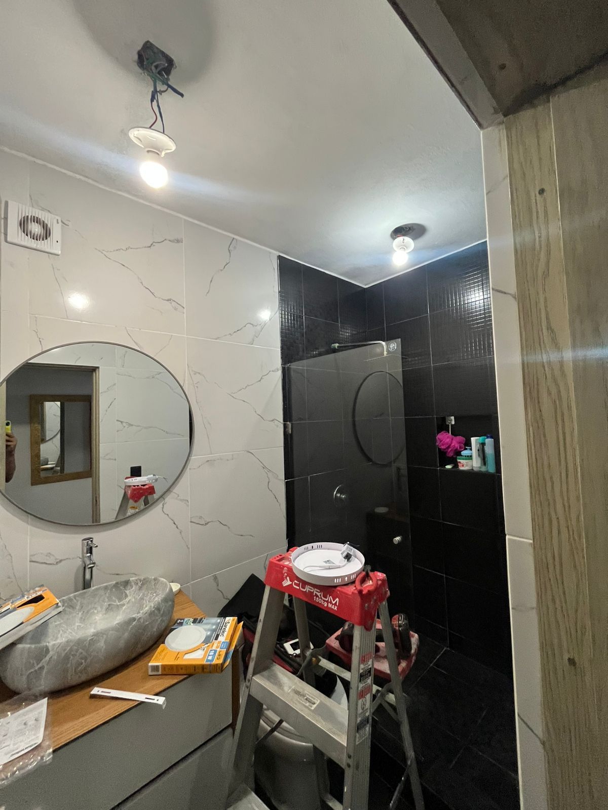 Un baño en renovación con azulejos blancos con estampado de mármol, pared de ducha negra, un espejo redondo y una escalera de mano.