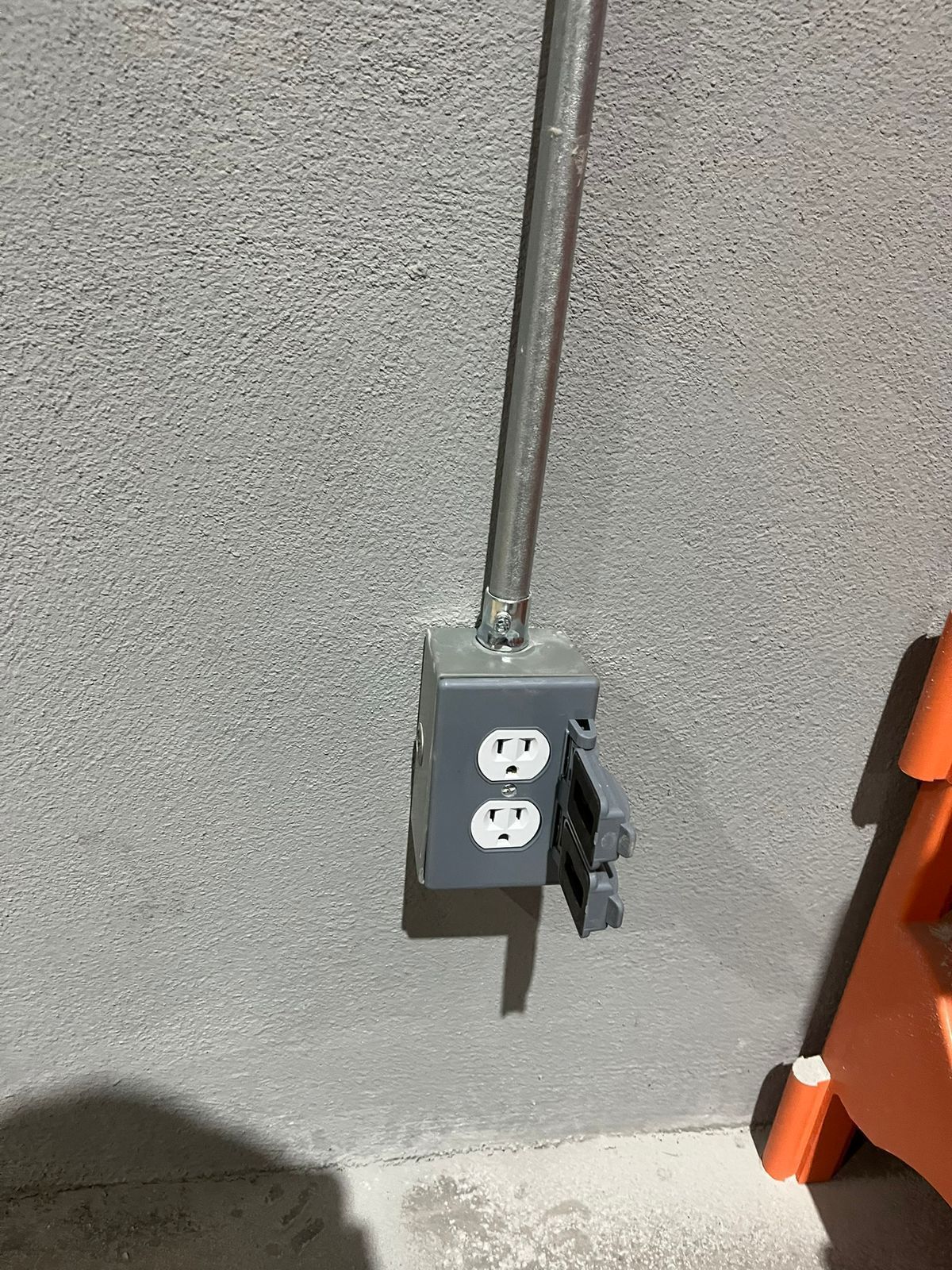 Un tomacorriente eléctrico gris, revestido de metal, montado en una pared exterior de estuco con una cubierta abierta resistente a la intemperie.