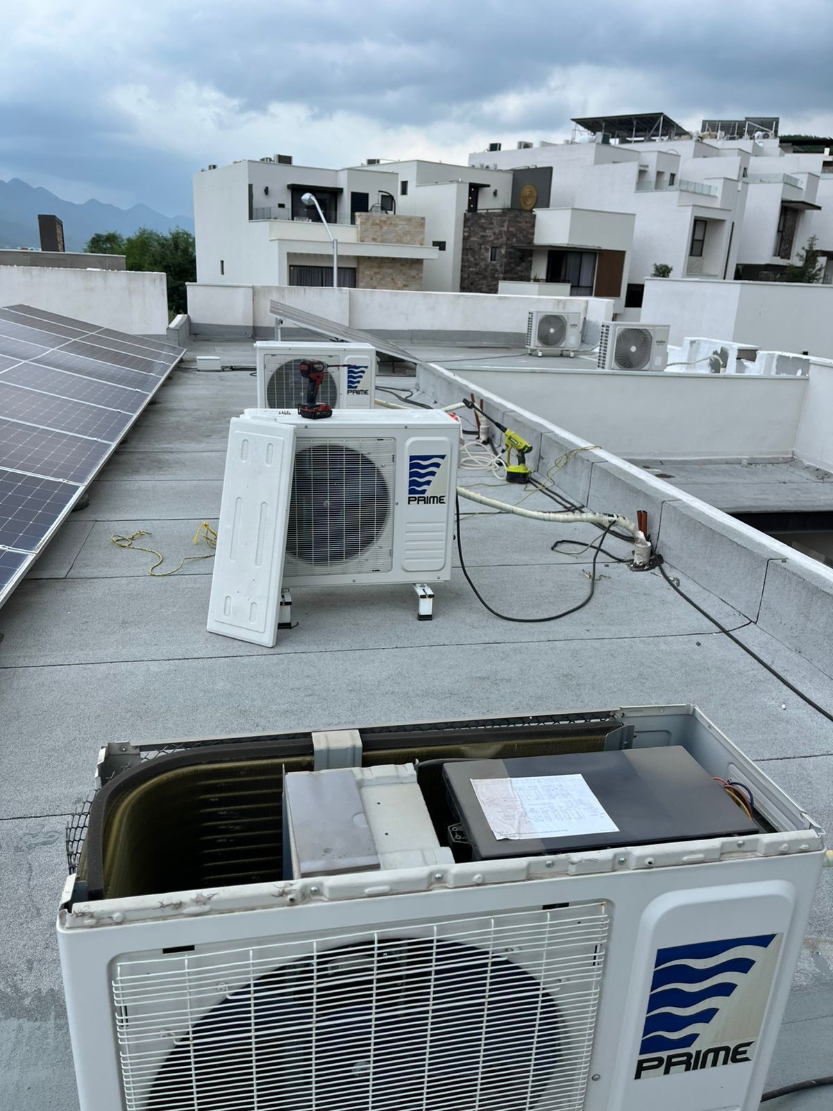 Tres unidades de aire acondicionado blancas colocadas sobre un techo plano cubierto de grava, con edificios residenciales al fondo.