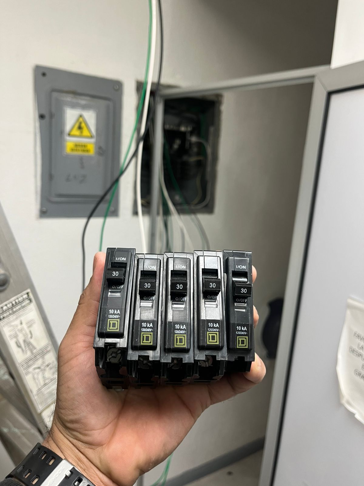 Una mano sostiene una fila de cinco disyuntores eléctricos delante de un panel eléctrico abierto montado en la pared.