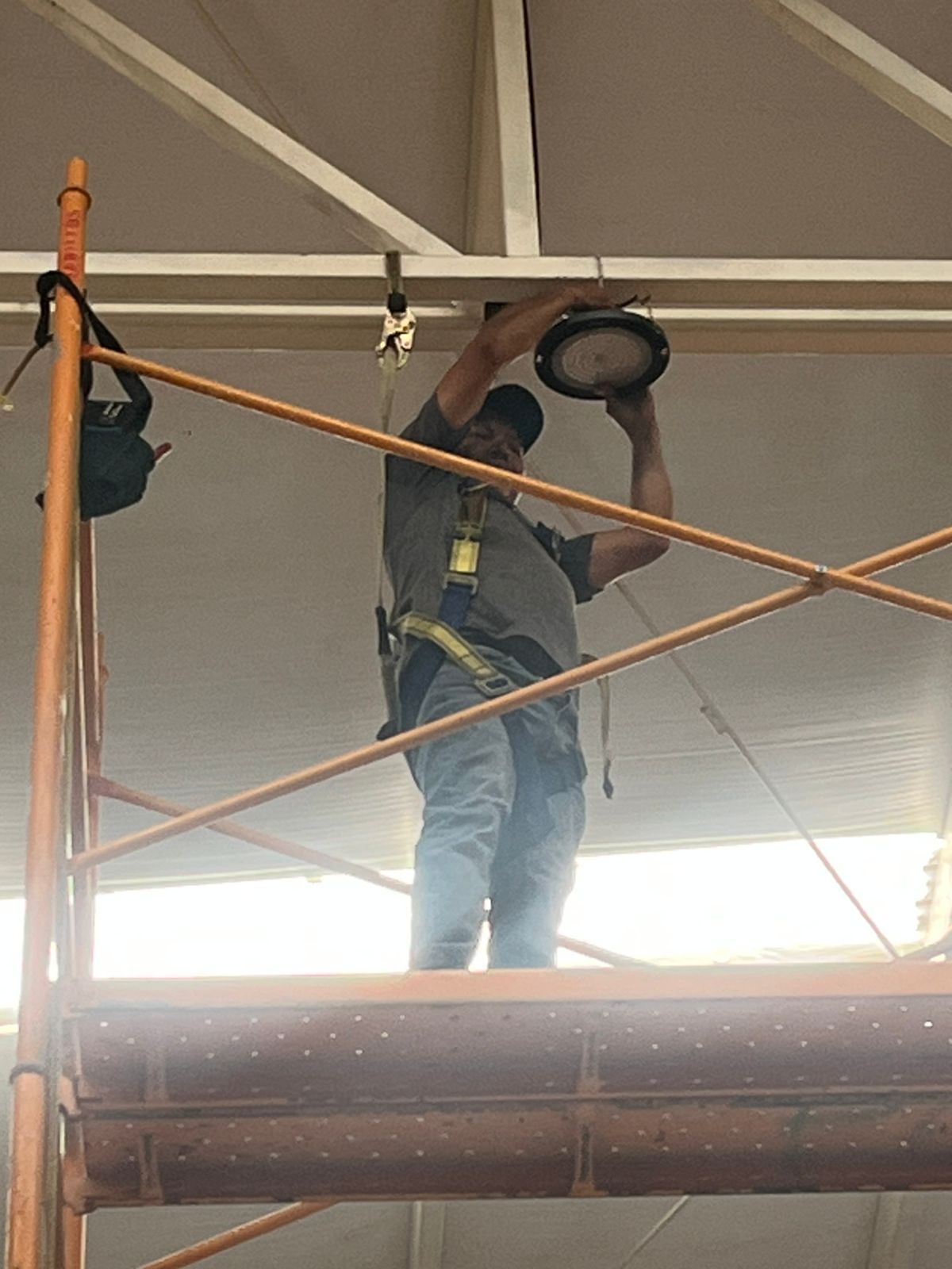 Una persona que lleva un arnés de seguridad se encuentra de pie sobre un andamio, trabajando en una lámpara de techo en un gran espacio interior.