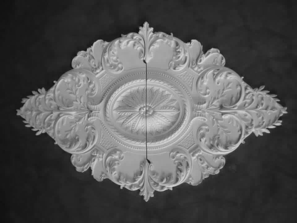 Wit, rond plafondmedaillon (rozet) met sierlijke reliëfdetails en een centraal bloemachtig ontwerp.