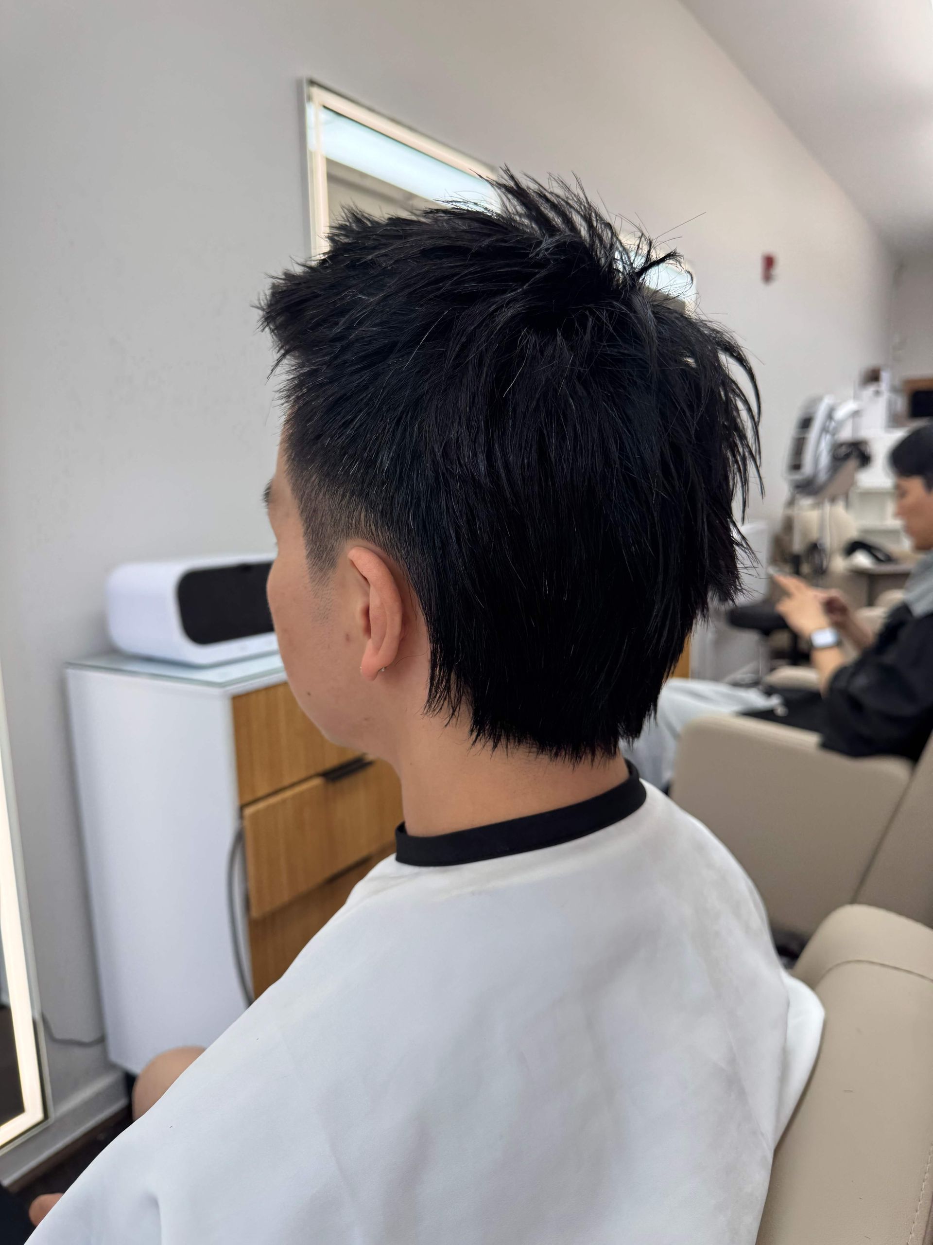 Person with short spiky hair sitting in a salon chair, seen from behind, with a mirror and stylist in the background