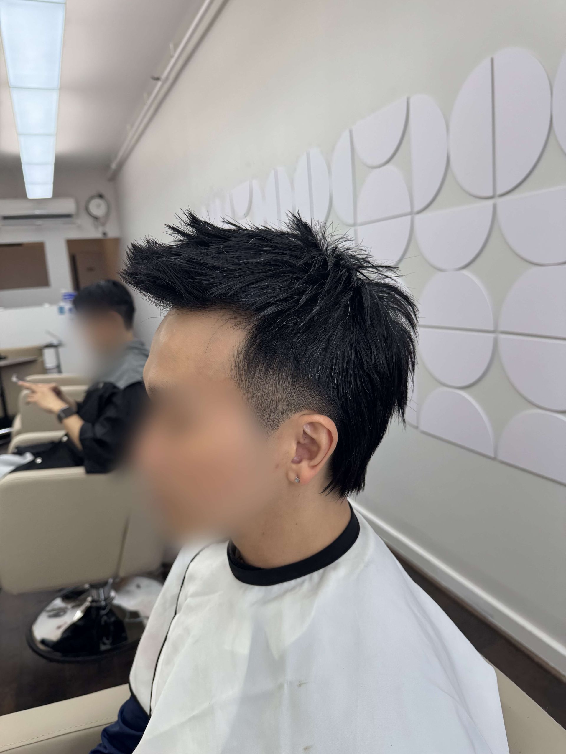Profile view of a person with spiky dark hair in a barber chair, wearing a white cape in a salon.