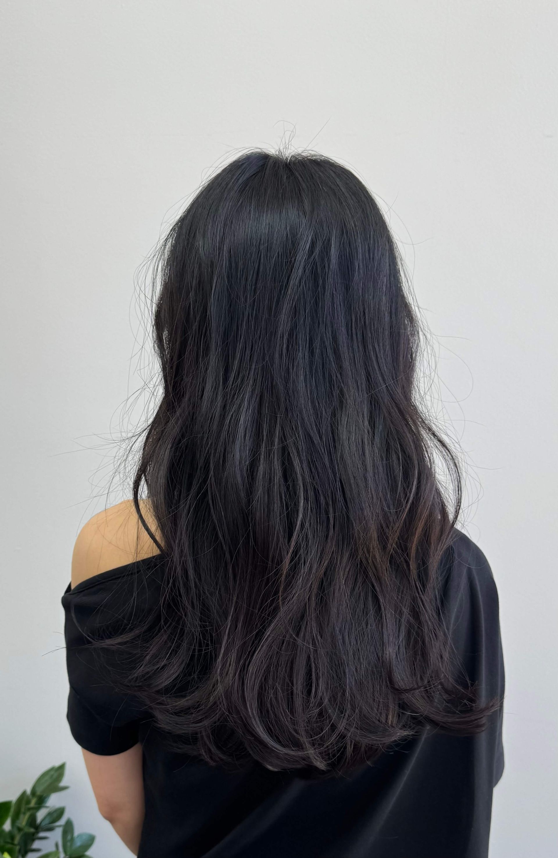 Person seen from behind with long dark hair, wearing a black off-shoulder top against a plain light wall.