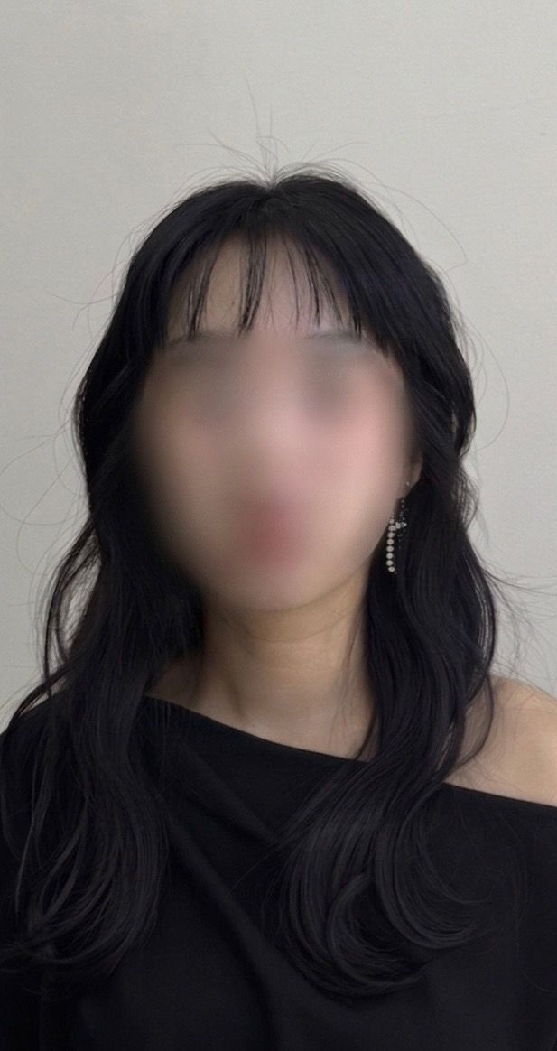 Portrait of a person with long dark hair wearing a black off-shoulder top against a plain wall