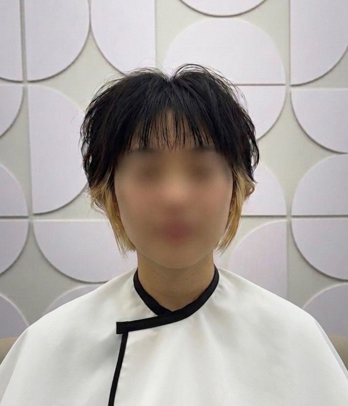 Person with short dark hair wearing a white top with black trim, standing against a patterned wall.