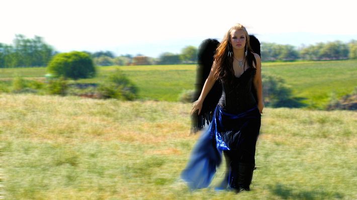 A woman in a black dress with black wings is walking through a field.