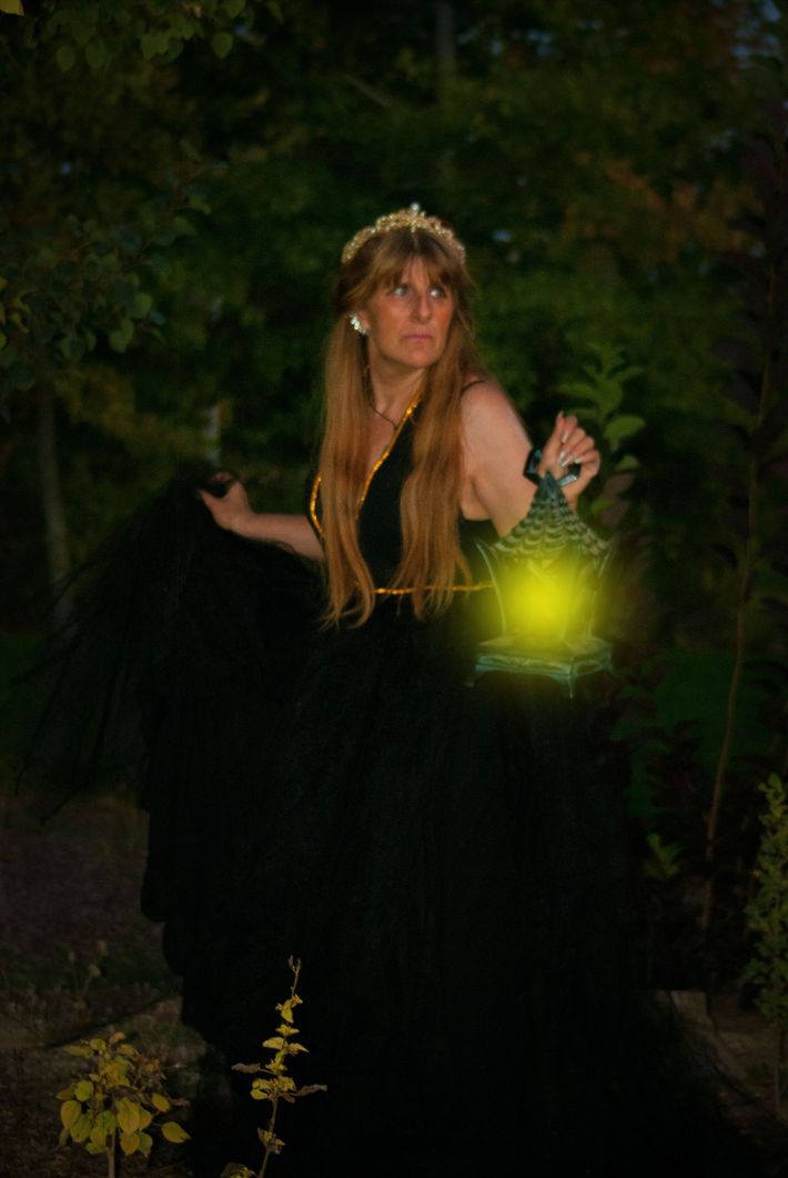 A woman in a black dress is holding a lantern in a dark forest.