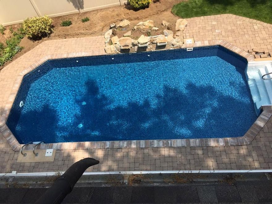 Backyard swimming pool with blue water, stone border, and patio chairs in the background