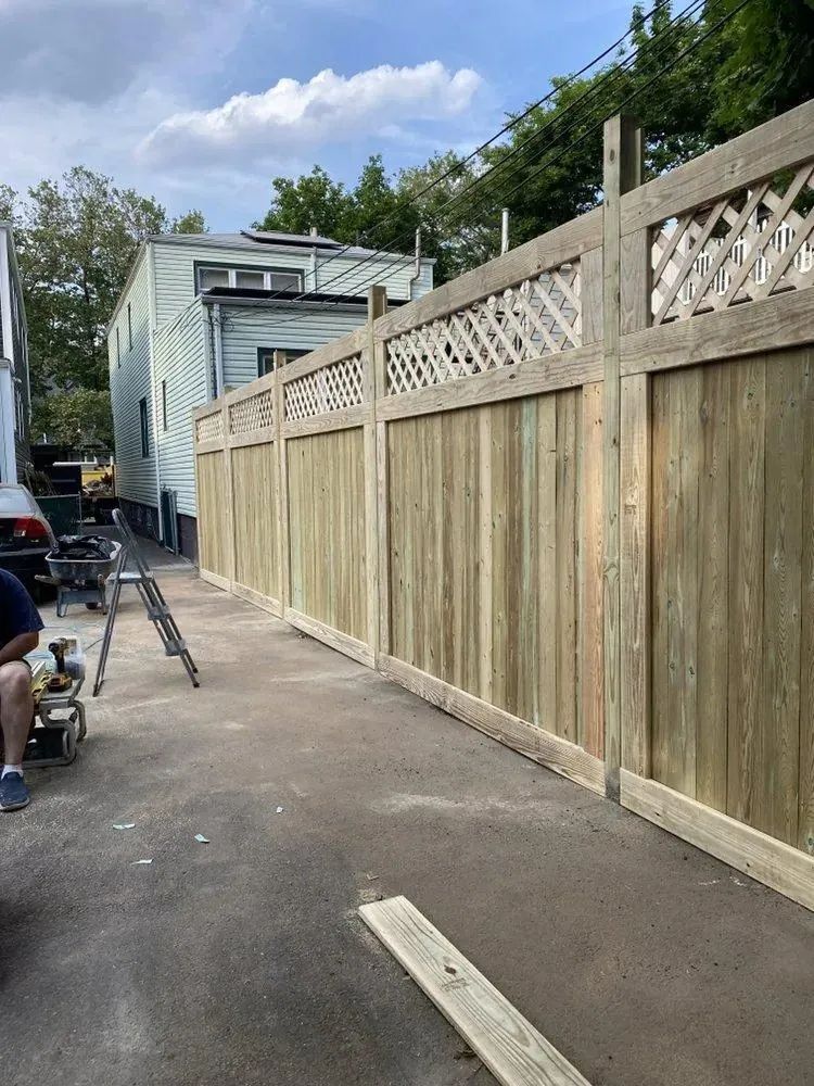 Long wooden fence along a driveway beside a white house under a partly cloudy sky
