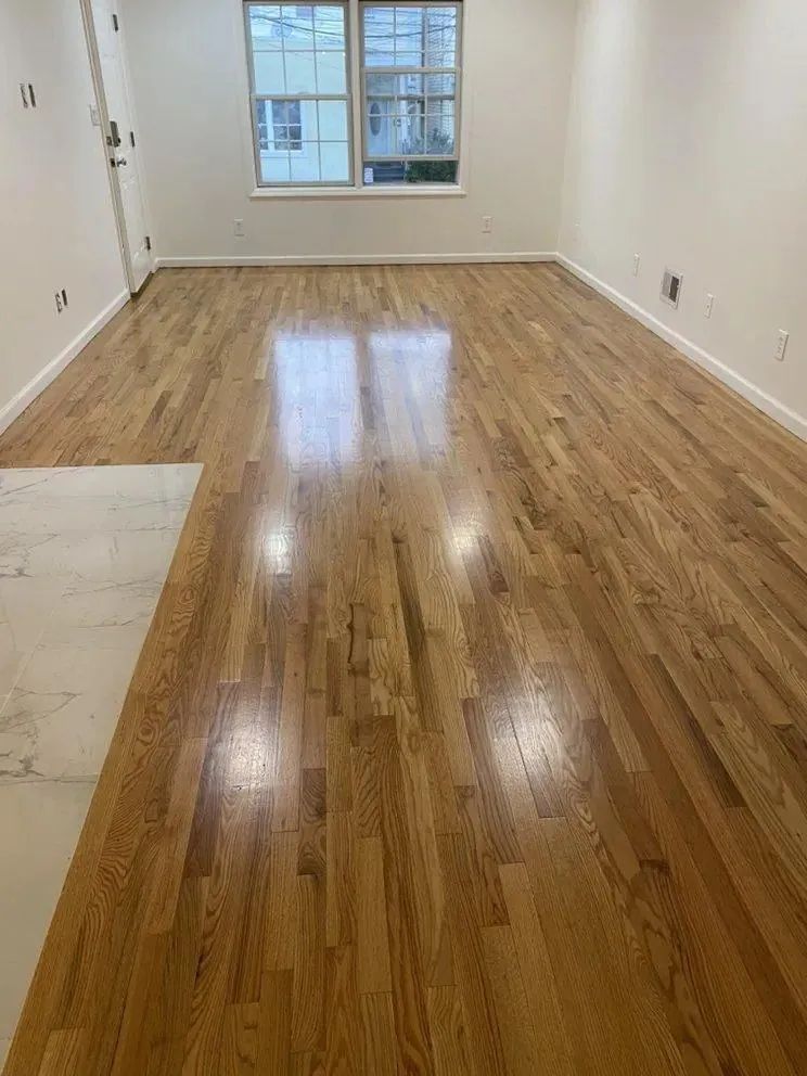 Empty room with glossy hardwood floors, white walls, and a window at the far end.