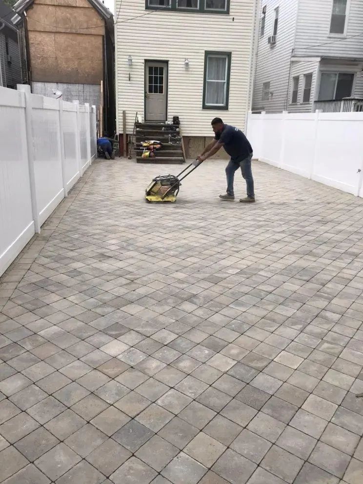 Person using a plate compactor on a paver driveway between white fences and a house.