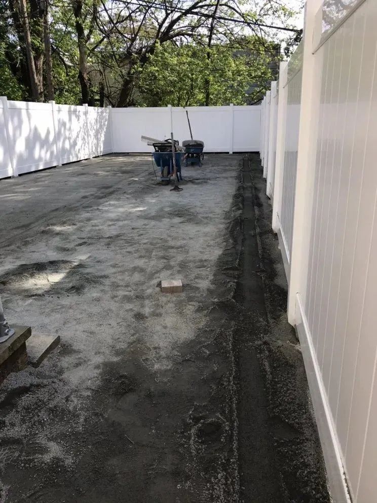 Long narrow outdoor yard with gravel, white fencing, trees, and a wheelbarrow near the center