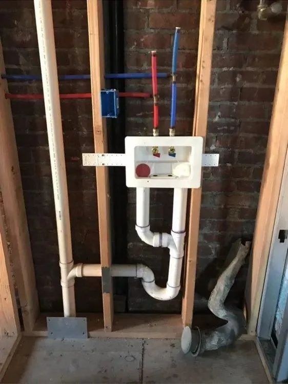 Exposed plumbing in unfinished wall framing with white access panel and pipes under a house.