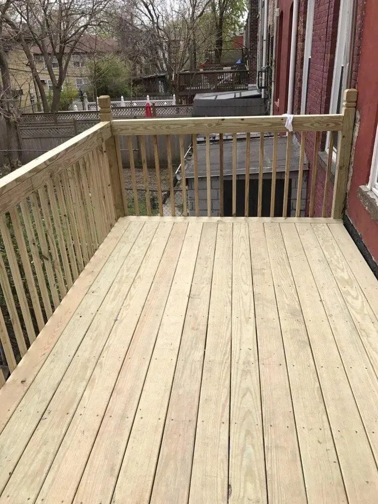 Wooden deck and railing beside a brick building, overlooking a backyard and parked car.