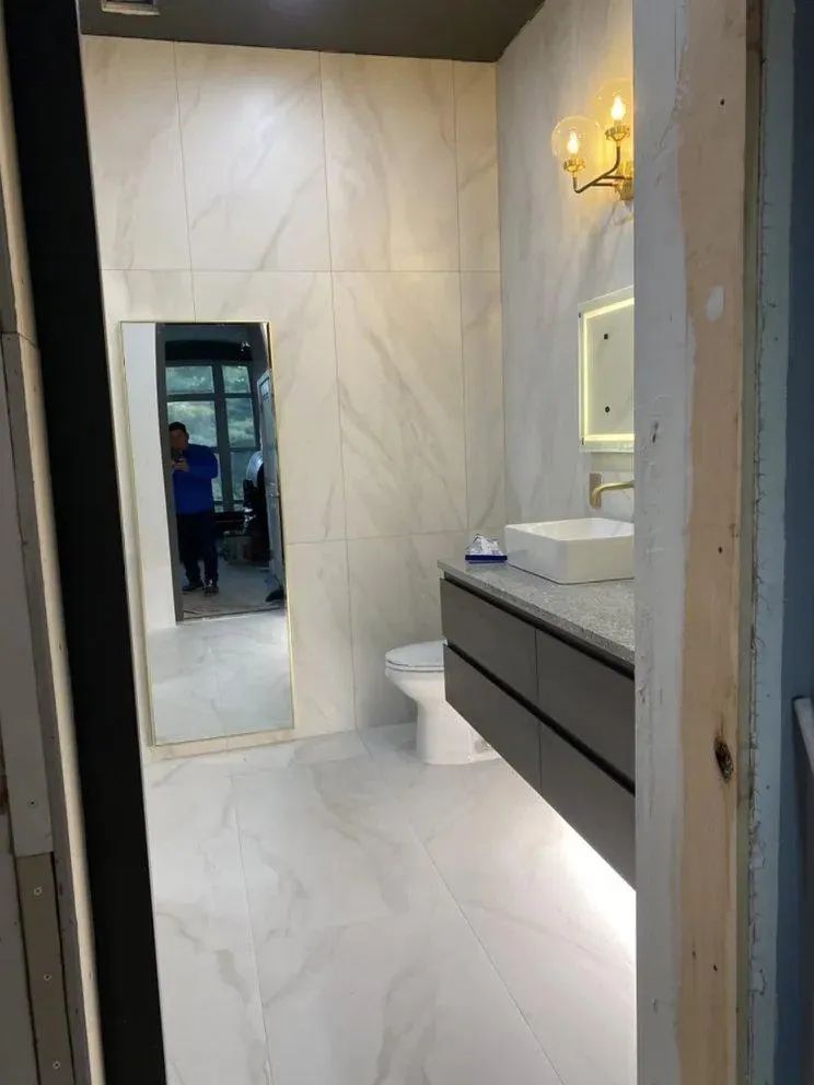 Small modern bathroom with white marble walls, vanity sink, mirror, and wall sconce lighting.