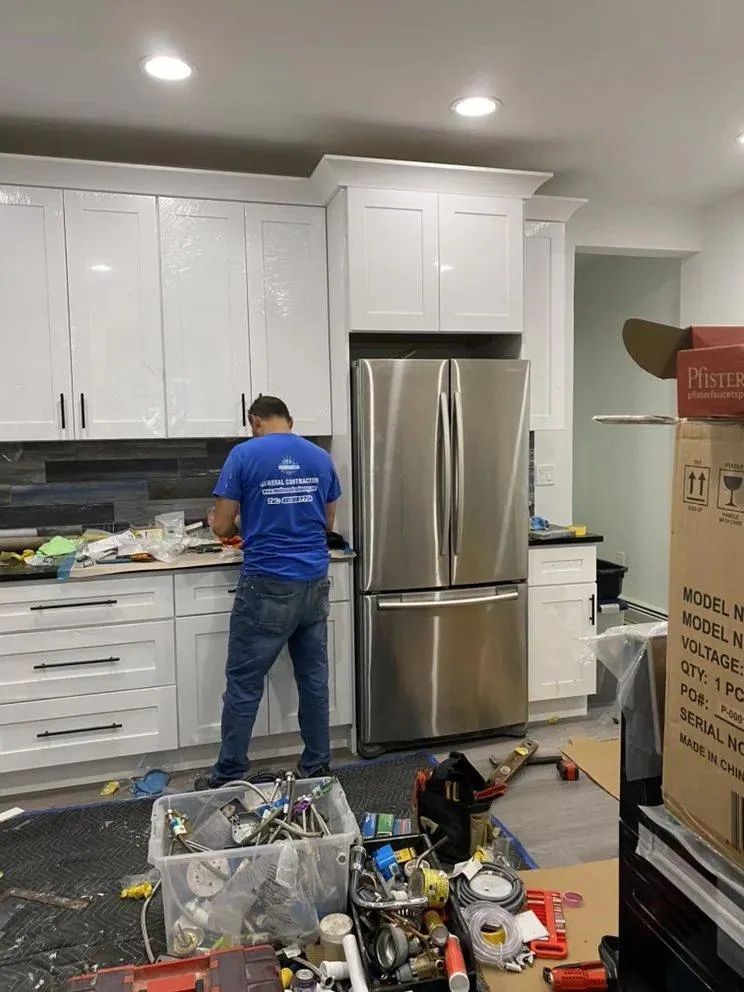 Person in blue shirt working in a kitchen with white cabinets, stainless steel fridge, and tools scattered on the floor