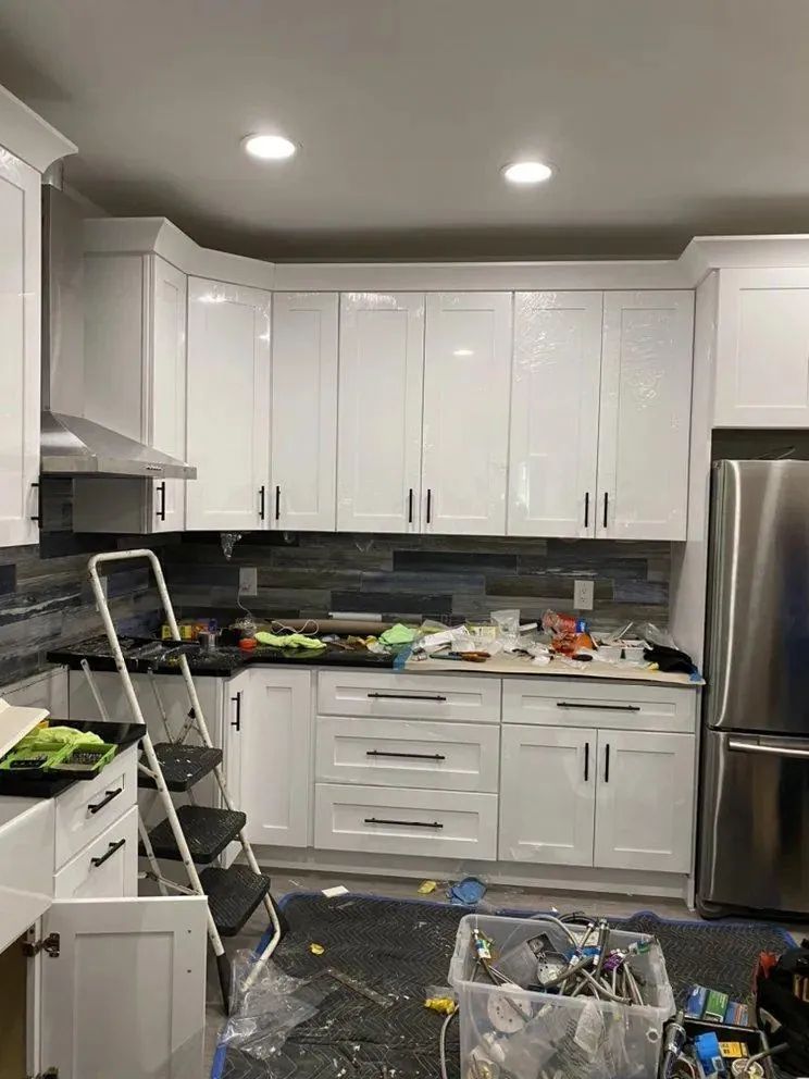 Messy white kitchen with cabinets, ladder, cleaning supplies, and clutter on floor and counters