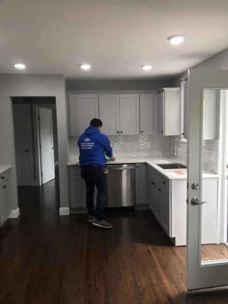 Person in blue jacket standing in a modern kitchen with white cabinets and dark wood floors.