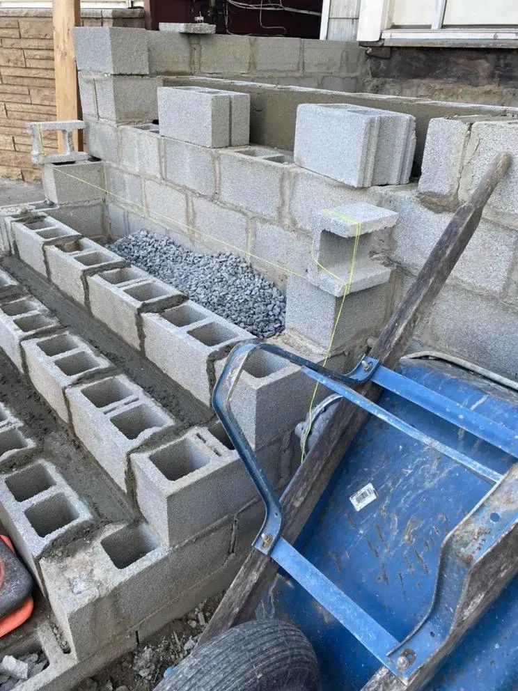 Partially built cinder block wall with loose blocks, gravel, and a blue wheelbarrow at a construction site