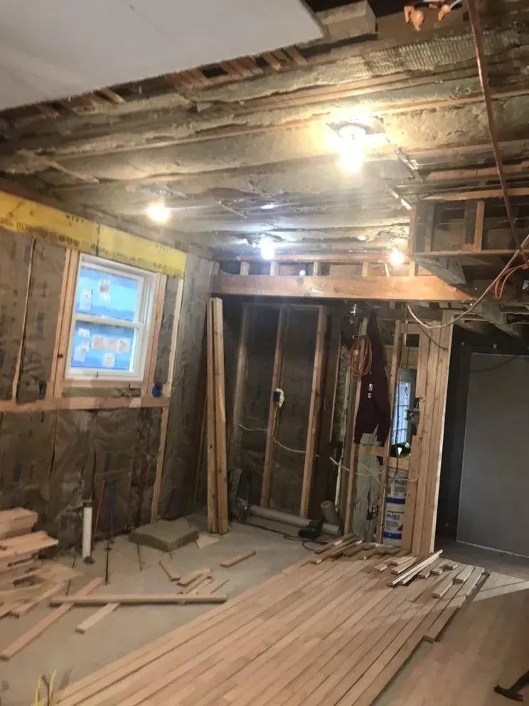 Renovation in progress in a gutted room with exposed studs, unfinished ceiling, and stacked lumber on the floor