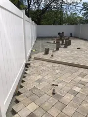 Empty patio with paver flooring, white fence, and several utility posts in a fenced yard